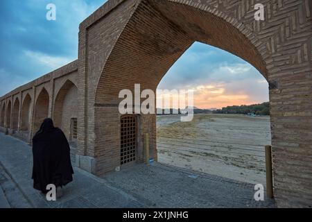 Die iranische Frau im schwarzen Tschador kommt an einem Bogen auf der Si-o-se-pol-Brücke vorbei, mit Blick auf das trockene Flussbett des Zayanderud während der Trockenzeit. Isfahan, Iran. Stockfoto