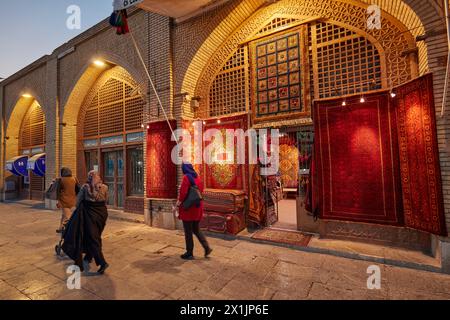 Die Leute gehen an einem Kunsthandwerksladen auf dem Platz Naqsh-e Jahan vorbei, wo eine Auswahl an handgefertigten persischen Teppichen in der Ladenfront ausgestellt wird. Isfahan, Iran. Stockfoto