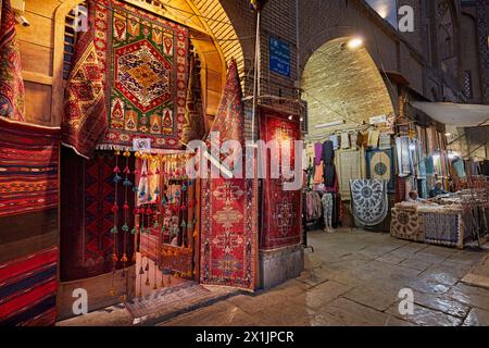 Eine Auswahl an handgefertigten persischen Teppichen, die am Eingang eines Kunsthandwerksgeschäfts auf dem bei Nacht beleuchteten Platz Naqsh-e Jahan ausgestellt werden. Isfahan, Iran. Stockfoto