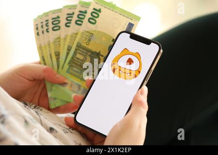 KIEW, UKRAINE - 1. APRIL 2024 Huya-Symbol auf dem Smartphone-Bildschirm und Geld in weiblicher Hand. IPhone-Display mit App-Logo und hundert Euro-Scheine in Mädchen Hand nah dran Stockfoto