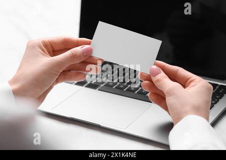 Frau mit Laptop, die leere Visitenkarte am weißen Tisch hält, Nahaufnahme. Leerzeichen für Text Stockfoto