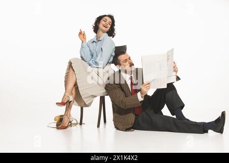 Glückliche, lächelnde elegante Frau, die auf einem Stuhl sitzt, ein Mann im klassischen Anzug sitzt auf dem Boden neben ihr und liest Tagebuch. Stockfoto