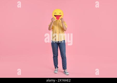 Frau, die das Gesicht mit Emoticon bedeckt und die Zunge auf rosa Hintergrund hinausragt Stockfoto