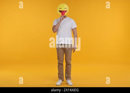 Mann, der das Gesicht mit Emoticon bedeckt, der die Zunge auf gelbem Hintergrund hinausragt Stockfoto