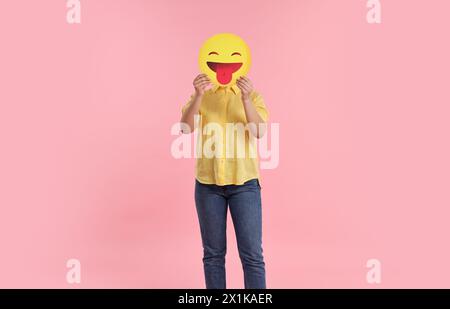 Frau, die das Gesicht mit Emoticon bedeckt und die Zunge auf rosa Hintergrund hinausragt Stockfoto