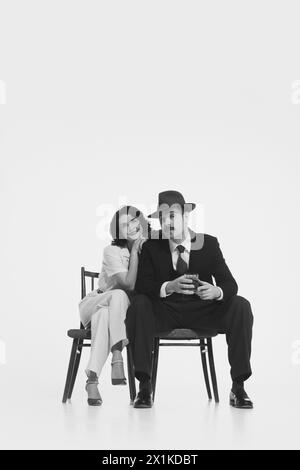 Einfarbiges Porträt eines jungen verheirateten Paares, eines Mannes mit Fedora-Hut und Anzug, der mit Whiskyglas sitzt, und einer eleganten lächelnden Frau, die sich auf die Schulter lehnt Stockfoto