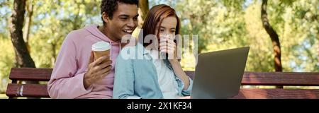 Ein vielseitiges Paar in lebendiger Kleidung, das auf der Parkbank sitzt und auf dem Laptop-Bildschirm vertieft ist. Stockfoto