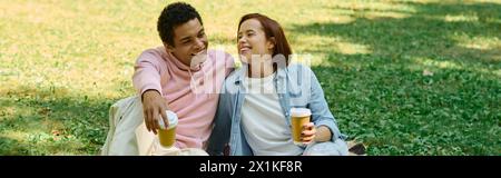 Ein ruhiger Moment, während ein vielseitiges, liebevolles Paar in lebendigen Kleidern zusammen auf dem Gras im Park sitzt. Stockfoto