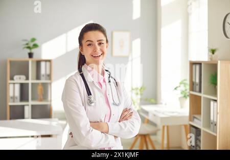Porträt einer jungen hübschen brünetten Ärztin in weißer medizinischer Uniform, die in ihrem Büro steht. Stockfoto