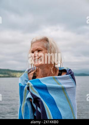 Ein Senior Open Water Wildschwimmer wärmt sich mit einem Handtuch nach einem erfrischenden kalten Wasser See Schwimmen - Frau schwimmen im Freien Gesundheit UK Stockfoto