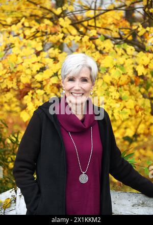 Happy Smile beleuchtet das Gesicht einer älteren Frau, während sie die gelben und goldenen Blätter des Herbstes genießt. Sie lehnt sich an einen rustikalen Holzzaun. Stockfoto