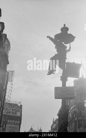 VE Day Scenes – rechts im Bild steht ein Mann auf einem Schild, der von einem Lampenpfosten aus in der Coventry Street ragt. Unter dem Mann befindet sich ein Schild, das die Fahrer zum Nuffield-Zentrum für H.M. Forces führt. Der untere Teil der Säule ist die Stütze für Ampeln. Links im Bild fliegt die Flagge der Vereinigten Staaten von einem Gebäude. Im linken Hintergrund befindet sich ein Horten für die Hollywoodkantine. Von einem Baum nach oben bis hinter die Kamera. Das Foto wurde von Walter Lassally aufgenommen, einem jungen Mann, der sechs Jahre zuvor als Flüchtling in Großbritannien angekommen war. Jahre später erklärte Walter das Stockfoto