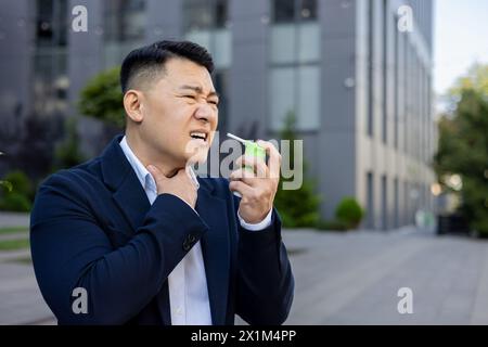Asiatischer junger Geschäftsmann, der in der Nähe des Arbeitsbüros draußen sitzt, Kehle mit der Hand hält, starke Schmerzen fühlt und medizinisches Spray verwendet. Stockfoto