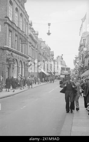 VE Day Scenes – im rechten Vordergrund laufen vier Männer auf dem Bürgersteig. Ein Mann hält das Revers seiner Jacke und der Mann neben ihm lächelt. Ein Doppeldeckerbus fährt entlang der Straße in Richtung der Kamera. Von einem Baum bis hinter die Kamera. Das Bild zeigt eine dichte Menschenmenge, die sich am 8. Mai 1945 - VE-Day vor den Toren des Buckingham Palace versammelt hat. Das Foto wurde von Walter Lassally aufgenommen, einem jungen Mann, der sechs Jahre zuvor als Flüchtling in Großbritannien angekommen war. Jahre später erklärte Walter, dass er als Kind in Berlin aufgewachsen sei, o Stockfoto