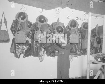 AIR RAID PRECAUTIONS IN KINGSTON HOUSE, KNIGHTSBRIDGE, LONDON, ENGLAND, C 1940 - Ein Mitglied des ARP-Personals überprüft die Anti-Gas-Kleidung und Ausrüstung der 61 Squad, die an einer Wand im Kingston House hängt. Die Stahlhelme, Gasmasken, Anti-Gas-Capes und wellington-Stiefel, die die Wand säumen, waren für den Einsatz bei einem Gasangriff unerlässlich. Beachten Sie auch die Reihen der gelagerten Ersatzhelme und Duty-Gas-Masken in Haversacks im rechten Vordergrund des Fotos. Das "R" auf seiner Tunika zeigt wahrscheinlich an, dass er von einer Rettungsaktion stammt. Stockfoto