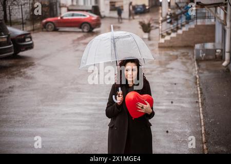 Eine Frau, die ein rotes Herz unter einem klaren Schirm hält. Der Schirm ist offen und die Frau steht im Regen. Stockfoto