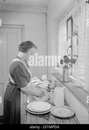 DIE COTTAGES VON FREEFOLK: DAS LEBEN IM DORF FREEFOLK, HAMPSHIRE, ENGLAND, Großbritannien, 1943 - Frau Shadwell steht am großen Waschbecken in der Küche ihres Hauses im Dorf Freefolk, Hampshire und wäscht das Geschirr. Das Waschbecken befindet sich unter dem Fenster mit einem großen Abtropfbrett auf beiden Seiten des Waschbeckens. Eine Vase voller Blumen ziert die Fensterbank, Stockfoto