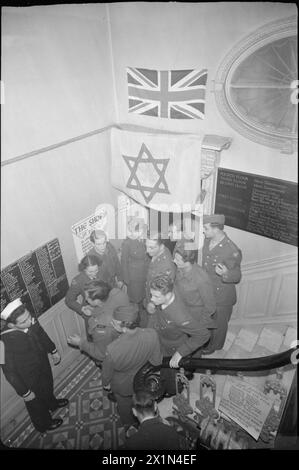 ALLIIERTE STREITKRÄFTE FEIERN JÜDISCHES NEUJAHR: RELIGIÖSE FEIERLICHKEITEN IM BALFOUR SERVICE CLUB, LONDON, Großbritannien, 1943 – Eine Gruppe alliierter Soldaten und Frauen drängen sich in den Flur des Balfour Service Club, einige stehen auf der Treppe, nach den Rosch-Hashanah-Feierlichkeiten. Auf den Tafeln links sind die Namen aller, die an dem fest teilnahmen, eingekreidet. Laut der Originalüberschrift konnten sich viele Menschen bei den Feierlichkeiten mit alten Freunden treffen. Eine Gewerkschaftsflagge und eine Flagge mit dem Davidstern hängen an der Wand über ihren Köpfen. Stockfoto