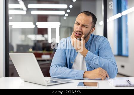 Junger hispanischer Mann, der im Büro arbeitet, am Schreibtisch sitzt und seinen Kiefer mit der Hand hält, grimmig ist und schwere Zahnschmerzen hat. Stockfoto
