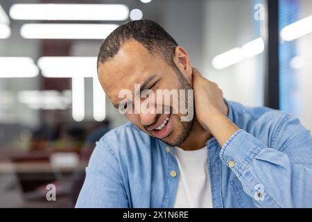 Nahaufnahme eines jungen hispanischen Mannes, der im Büro arbeitet, am Arbeitsplatz sitzt und seine Hand um den Hals hält und unter Schmerzen leidet. Stockfoto