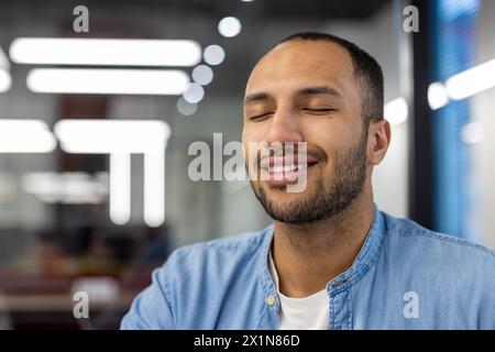 Nahaufnahme eines ruhigen und lächelnden jungen arabischen Mannes, der im Arbeitsbüro sitzt und mit geschlossenen Augen träumt. Stockfoto