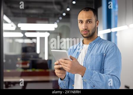 Porträt eines ernsten jungen lateinamerikanischen Mannes, der im Büro steht, ein Tablet hält und in die Kamera blickt. Stockfoto