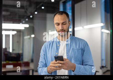 Ernster junger hispanischer Mann in Denim-Hemd, der im modernen Büro steht und das Handy benutzt. Stockfoto