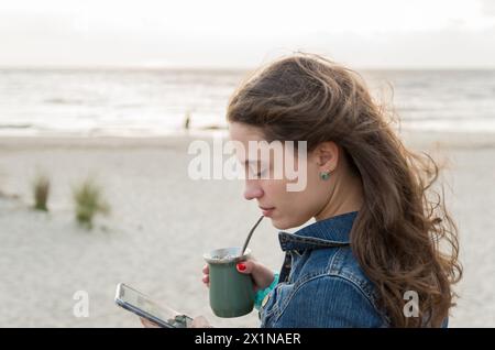 Schöne junge Frau trinkt Chimarao, Mate (Yerba Mate Infusionsgetränk) am Strand in Uruguay. Stockfoto