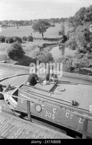 BRITISH CANALS IN WARTIME: TRANSPORT IN BRITAIN, 1944 – Ein Kanalboot, das in Rickmansworth registriert ist und wahrscheinlich zur Familie Gribbis gehört, fährt in der Sonne über das Aquädukt, das den Grand Union Canal über den Fluss Nene bei Wolverton in Buckinghamshire führt. Der Bootsmann, wahrscheinlich Mr. Gribbis, kann gesehen werden, wie er das Boot über das Aquädukt steuert und den Blick auf die Landschaft bewundert. Unter ihm kann man einen Mann sehen, der am Fluss Nene fischt. Stockfoto