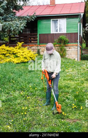 Frau schneidet Gras mit einem Fadenschneider in ihrem ländlichen Holzhaus. Rasenpflege im Garten im Frühjahr Stockfoto