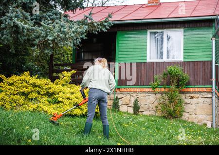 Frau schneidet Gras mit einem Fadenschneider in ihrem ländlichen Holzhaus. Rasenpflege im Garten im Frühjahr Stockfoto