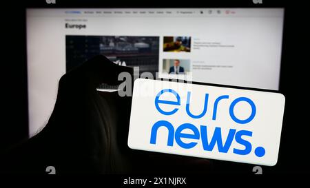 Person, die ein Mobiltelefon mit dem Logo des belgischen Nachrichtenunternehmens Euronews SA vor der Website des Unternehmens hält. Schwerpunkt auf dem Display des Telefons. Stockfoto