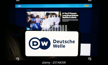 Person mit Mobiltelefon mit Logo der Deutschen Welle (DW) vor der Webseite. Schwerpunkt auf dem Display des Telefons. Stockfoto