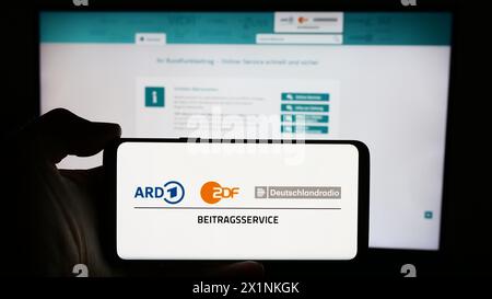 Person mit Handy mit Logo des ARD ZDF Deutschlandradio Beitragsservice vor der Webseite. Schwerpunkt auf dem Display des Telefons. Stockfoto