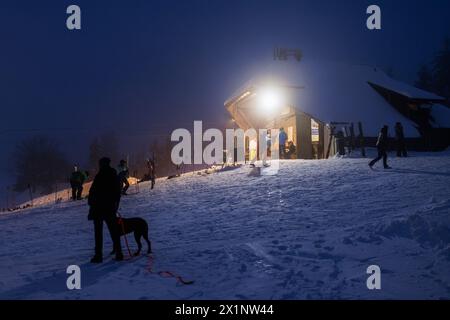 St. Peter, Deutschland. April 2024. Eine Frau steht mit ihrem Hund vor dem hell beleuchteten Kaibenlochlift-Gebäude. Ein Snowboarder fährt die Piste am Kaibenloch Lift hinunter. Die Wintersportsaison im Schwarzwald endete vor Wochen. Nach sommerlichen Temperaturen auf dem Schwarzwaldgipfel der Kandel kehrte Mitte April der Winter zurück - mit viel Neuschnee. Ab Mittwochabend öffnen die Kandellifte und bieten Flutlichtantrieb für Skifahrer und Snowboarder. Quelle: Philipp von Ditfurth/dpa/Alamy Live News Stockfoto