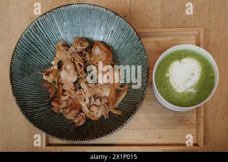 Schweinebraten mit Gemüse, bestreut mit Sesamsamen, gepaart mit einer cremigen Matcha-Grüntee-Latte, kunstvoll mit einem herzförmigen Schaum garniert, Stockfoto