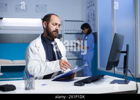 Bild zeigt einen engagierten kaukasier Arzt, der seine Notizen am Schreibtisch der Klinik überprüft und analysiert. Junger Mann mit weißem Laborkittel, der sich auf die ärztliche Behandlung mit Patienten vorbereitet. Stockfoto