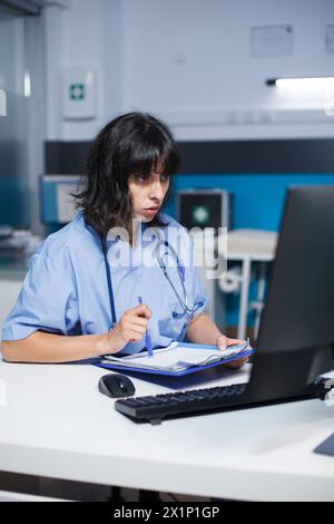 Eine fokussierte Krankenschwester untersucht und liest ihre Notizen im Büro der Klinik neu. Bei der Vorbereitung auf die medizinischen Konsultationen verwendet der kaukasische Arzt den Desktop-Monitor und das Clipboard. Stockfoto