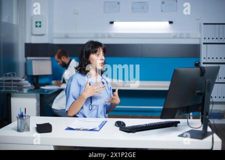 Eine Krankenschwester in einem Krankenhaus kommuniziert über einen Videoanruf mit ihrem Desktop-pc. Kaukasische Frau saß im Klinikbüro und hatte ein virtuelles Meeting. Zusammenarbeit und Technologie im Gesundheitswesen. Stockfoto