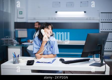 Lächelnde Praktizierende, die am Schreibtisch des Klinikbüros sitzt, spricht im Festnetz. Kaukasische Krankenschwester telefoniert mit einem Desktop-Computer in einem Krankenhauszimmer. Stockfoto
