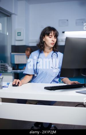 Im Klinikbüro, engagierter Arzt, der die Patientenakten mit dem Computer analysiert. Krankenschwester überprüft Patientendaten auf einem Desktop-Computer im Krankenhaus mit besonderer Aufmerksamkeit. Stockfoto