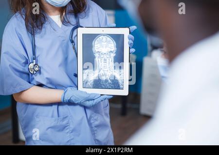 In der medizinischen Praxis verwendet der Arzt Tablette, um einen Scan des Schädels eines Patienten zu analysieren. Selektive Fokussierung der Schwester in blauen Peelings, die ein Gerät mit Röntgenbild halten. Fortschrittliche Technologie und fachkundige Betreuung. Stockfoto