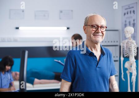 Älterer Mann mit Brille in einer Physiotherapieeinrichtung. Pensionierter Senior Patient lächelt in die Kamera, als er sich auf seine Genesung in der Klinik für Rehabilitationsbehandlung durch Fitness vorbereitet. Stockfoto