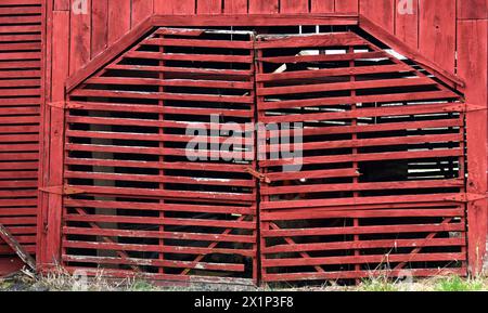 Rote Holzscheune hat geometrisch gerahmte Doppeltüren. Es sind Holzlatten, die gebrochene Bretter fehlen. Stockfoto