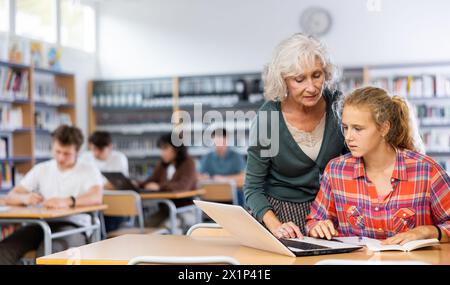 Eine freundliche Lehrerin hilft Schülerinnen, Informationen auf einem Laptop in der Schulbibliothek zu finden Stockfoto