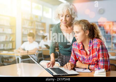 Eine freundliche Lehrerin hilft Schülerinnen, Informationen auf einem Laptop in der Schulbibliothek zu finden Stockfoto
