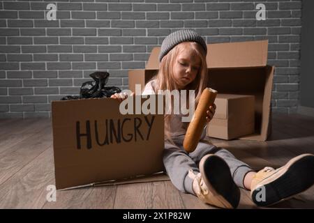 Obdachloses kleines Mädchen mit Brot, das um Essen bettelt, während es auf dem Boden sitzt Stockfoto