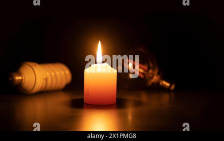 Blackout-Konzept, Eine weiße brennende Kerze in Nahaufnahme auf einer schwarzen Oberfläche vor zwei Glühbirnen auf schwarzem Hintergrund Stockfoto