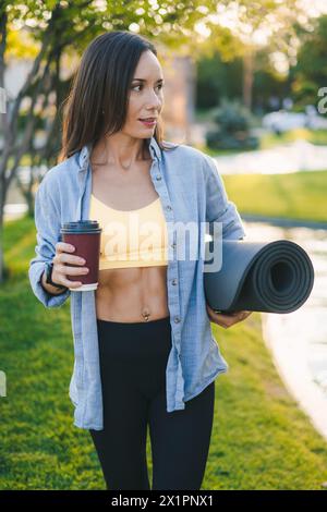 Erwachsene schöne lächelnde Frau mit einer Tasse Kaffee und einer Rollmatte, stehend im Park. Machen Sie eine Pause. Stockfoto