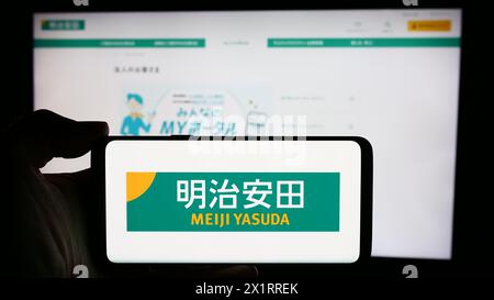 Person, die ein Mobiltelefon mit dem Logo des japanischen Unternehmens Meiji Yasuda Life Insurance Company vor der Webseite hält. Schwerpunkt auf dem Display des Telefons. Stockfoto